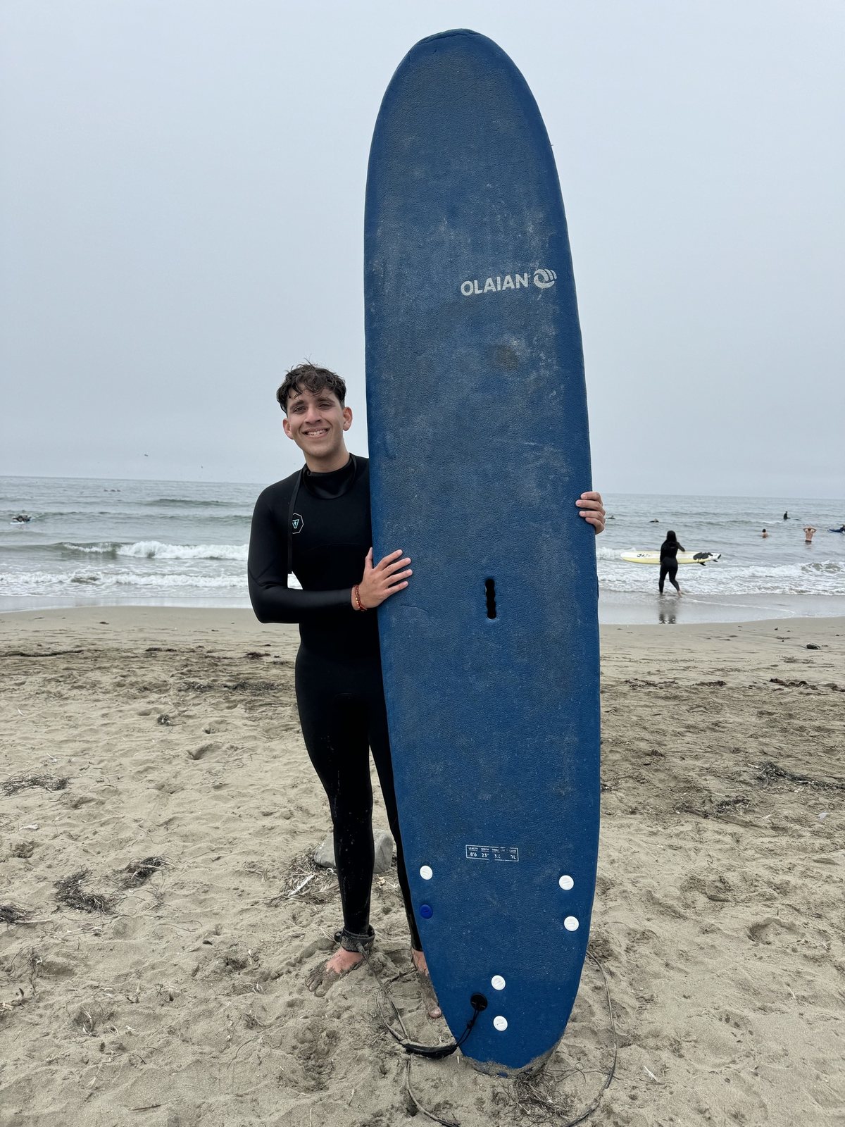 Aakash on the beach holding a blue soft-top surfboard